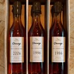 Trois bouteilles de la Collection Unique Armagnacs Darroze millésimes 2006, 1996 et 1986 dans une boîte en carton, présentées sur une surface en bois.